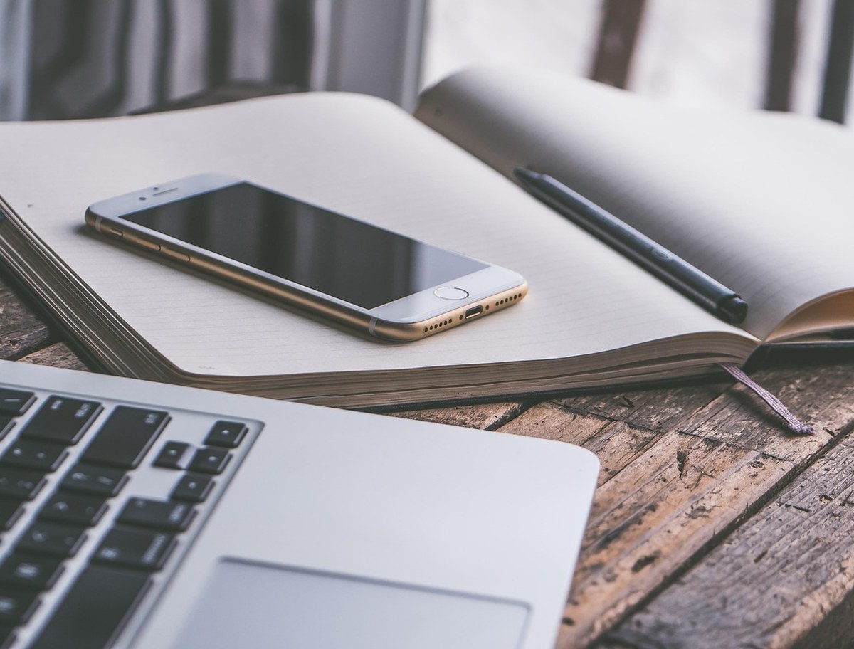 MacBook mit aufgeschlagenem Notizbuch und Smartphone auf hellem Holztisch